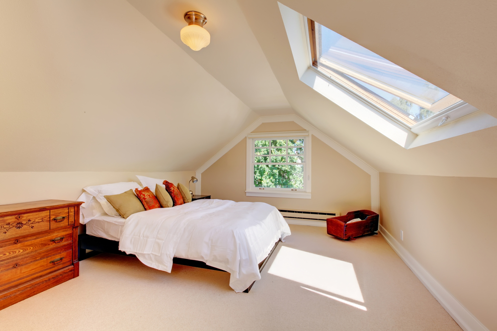 A cozy attic bedroom with a bed, white bedding, colorful pillows, a wooden chest of drawers, a skylight, and a small window letting in sunlight. A wooden chest sits near the window on a beige carpeted floor.