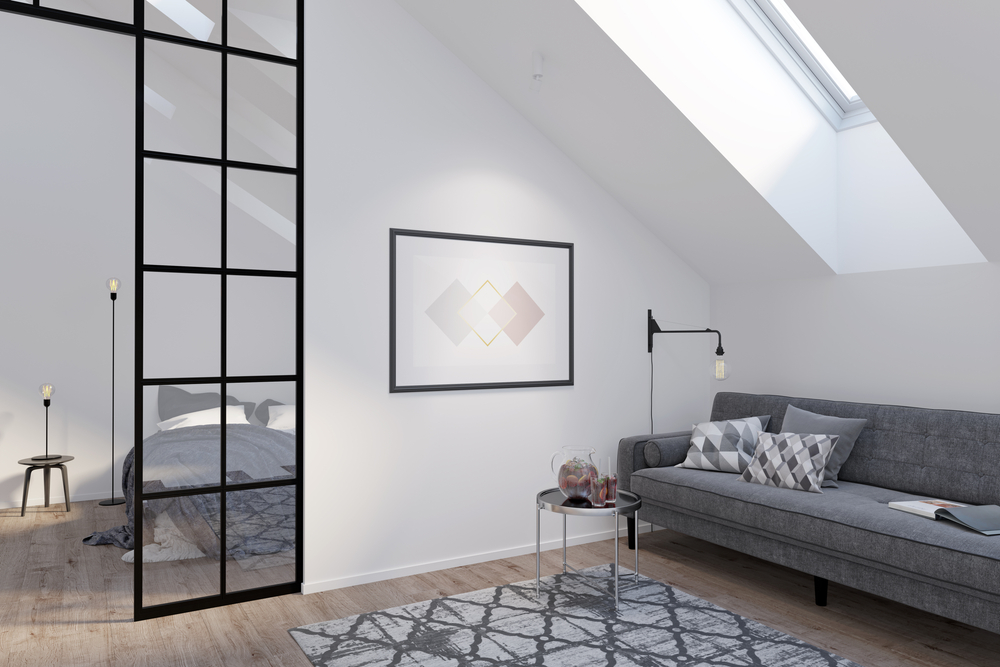 A modern loft with a skylight, gray sofa, patterned rug, side table, and wall art. A glass partition separates the living area from a bedroom with a bed and nightstand. The space is bright and minimally decorated.