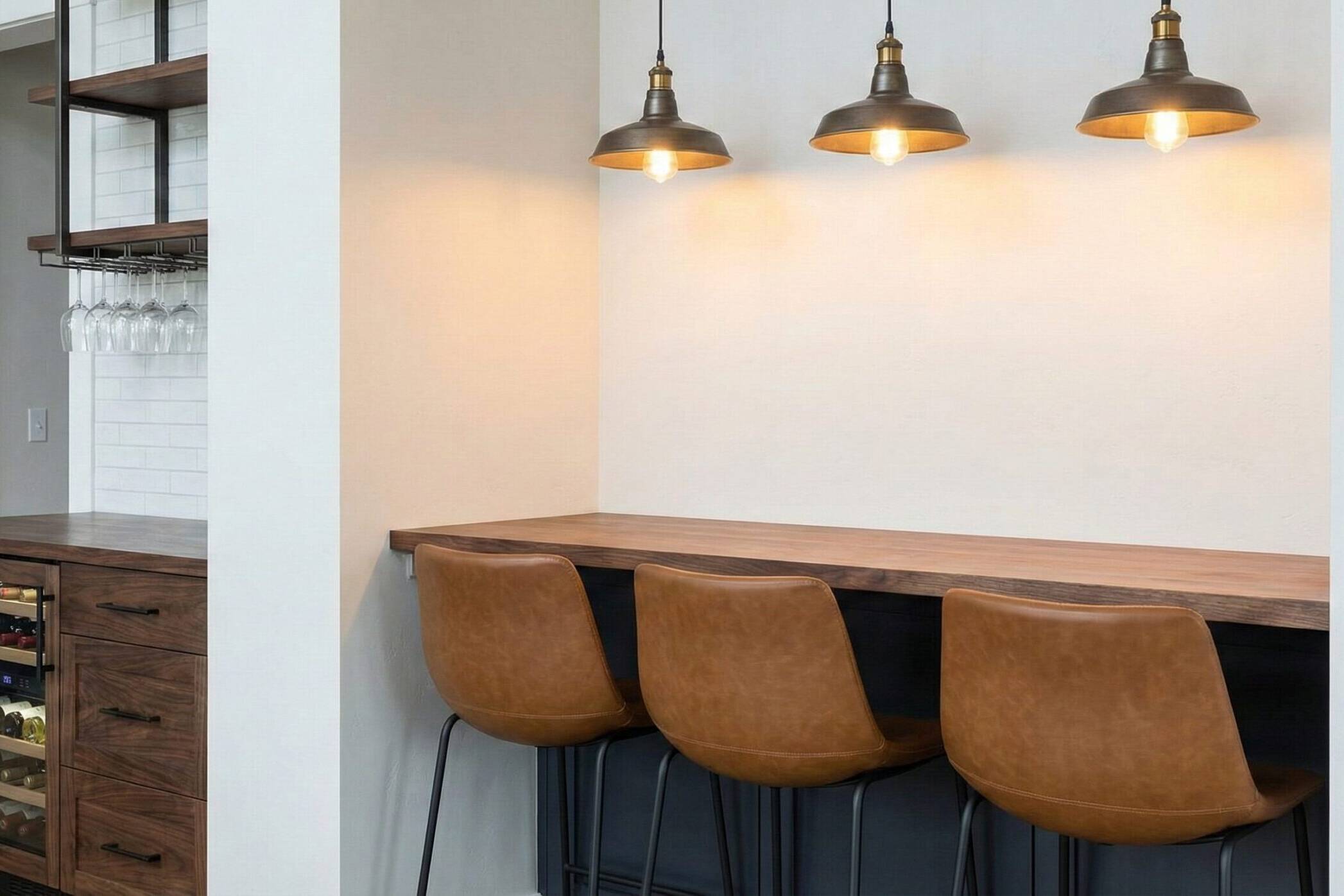 A modern bar area with three brown leather bar stools, a wooden counter, and three hanging pendant lights. Adjacent is a built-in wine fridge, glassware, and shelves with a white backsplash.