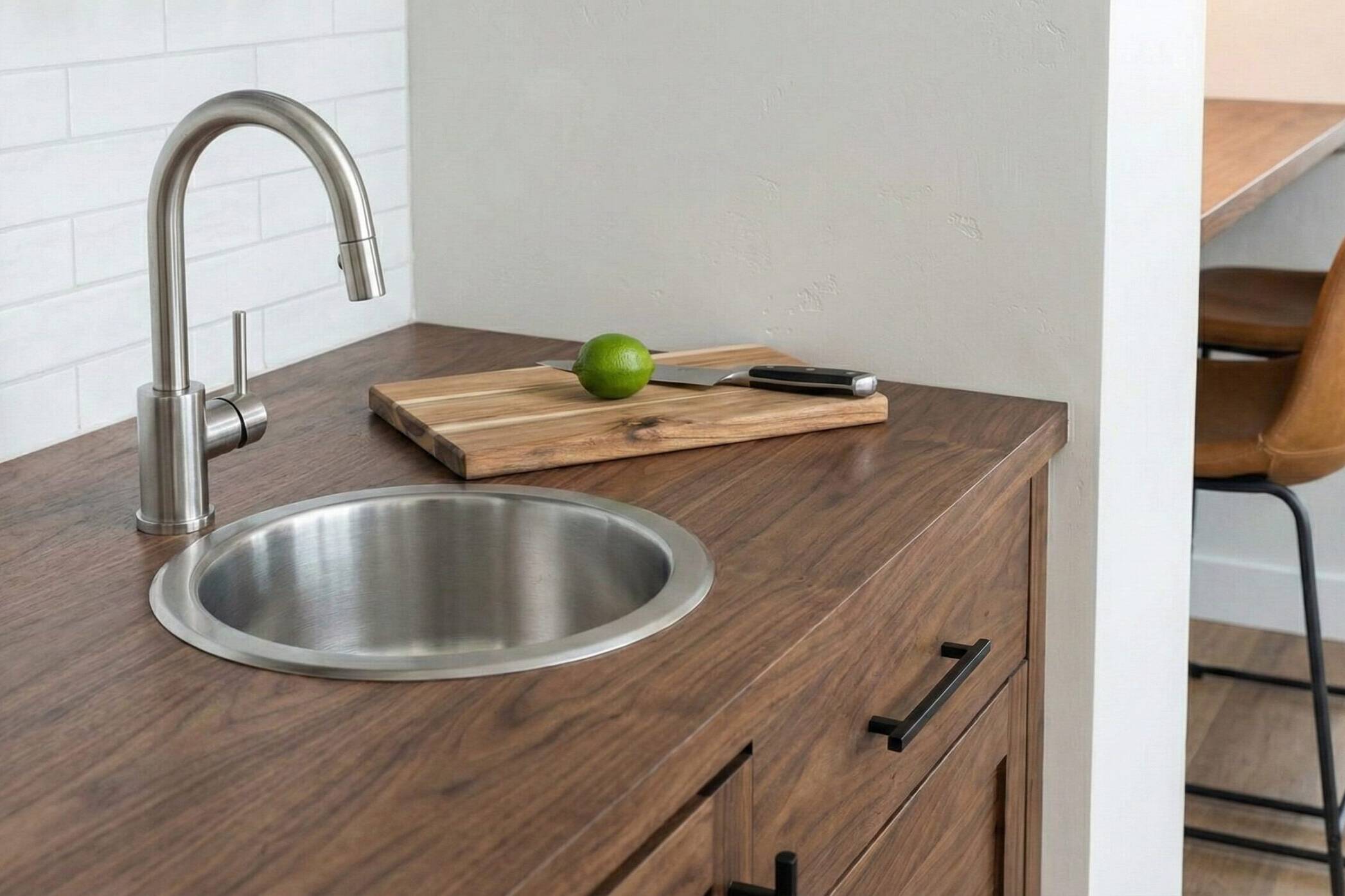 A modern kitchen countertop with a round stainless steel sink, a curved faucet, a wooden cutting board, a whole lime, and a knife. Two brown chairs and a wooden table are visible in the background.