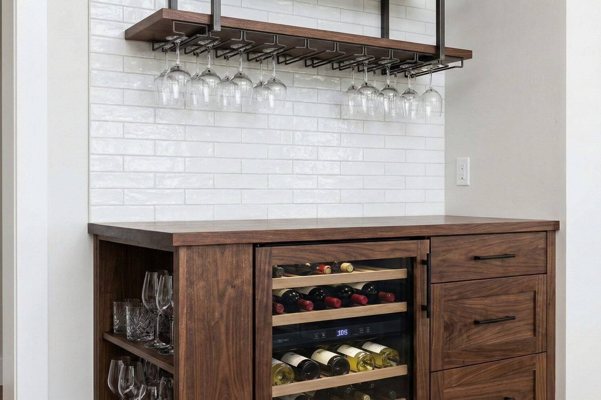 A wooden home bar with a wine fridge, wine bottles, glass shelves, four drawers, and an overhead rack holding hanging wine glasses. The backsplash features white subway tiles.
