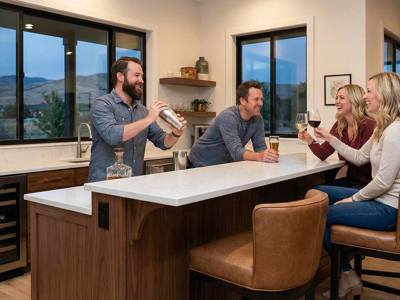 Four people are gathered in a modern kitchen. A man is shaking a cocktail shaker behind the counter while the others, holding drinks, smile and chat. Large windows show a scenic outdoor view.