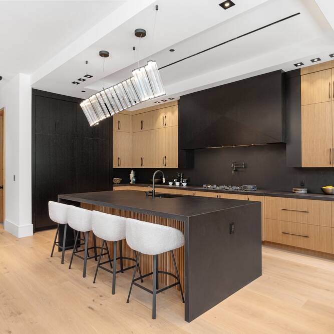 Modern kitchen with light wood floors, black countertops and backsplash, natural wood cabinets, a large island with four upholstered stools, and a contemporary rectangular pendant light above the island.