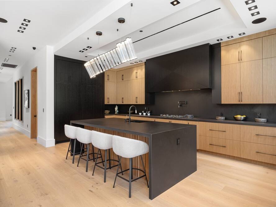 Modern kitchen with light wood floors, black countertops and backsplash, natural wood cabinets, a large island with four upholstered stools, and a contemporary rectangular pendant light above the island.