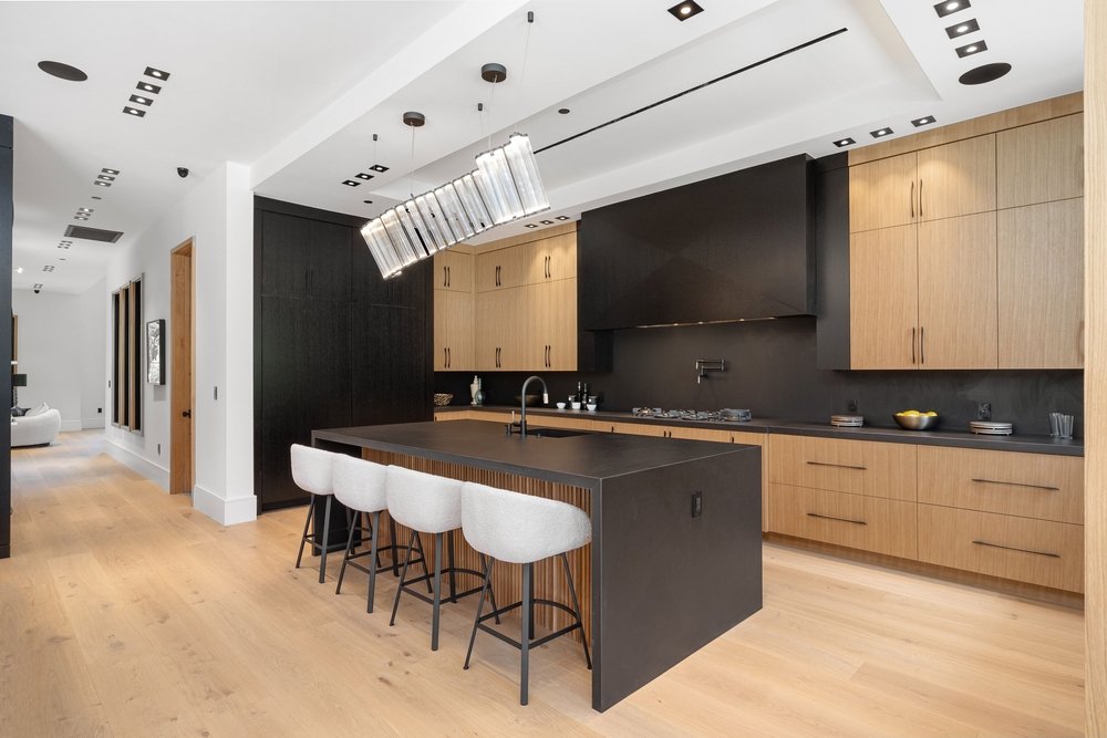 Modern kitchen with light wood floors, black countertops and backsplash, natural wood cabinets, a large island with four upholstered stools, and a contemporary rectangular pendant light above the island.