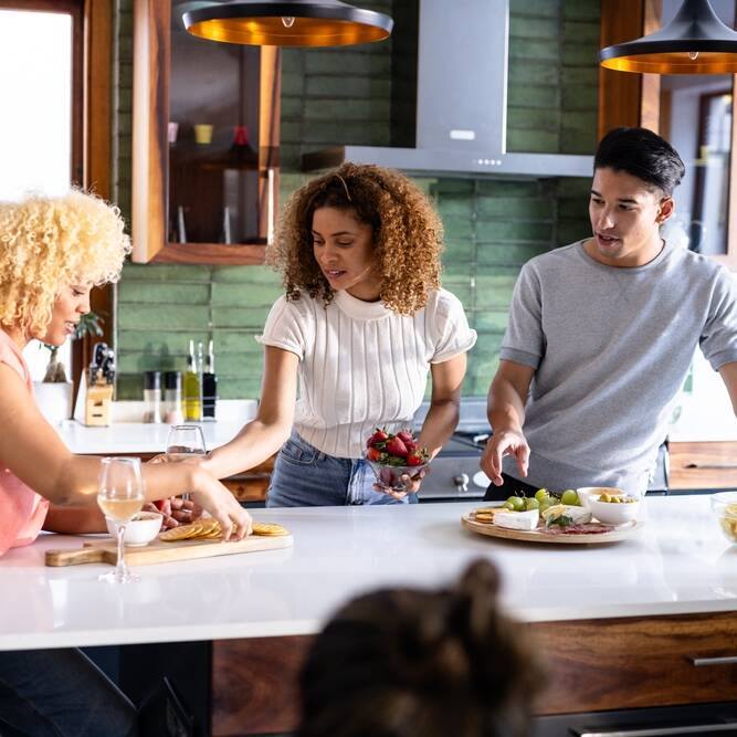 Three people gather around a kitchen island, preparing and sharing snacks and drinks, while chatting. A fourth person, mostly out of frame, sits nearby. The kitchen has green walls and modern decor.