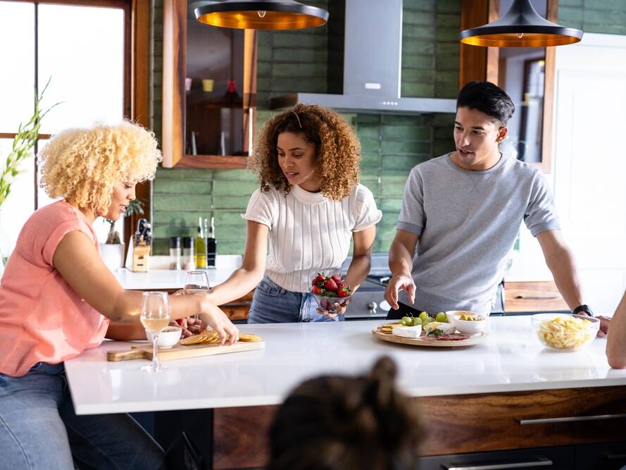 Three people gather around a kitchen island, preparing and sharing snacks and drinks, while chatting. A fourth person, mostly out of frame, sits nearby. The kitchen has green walls and modern decor.