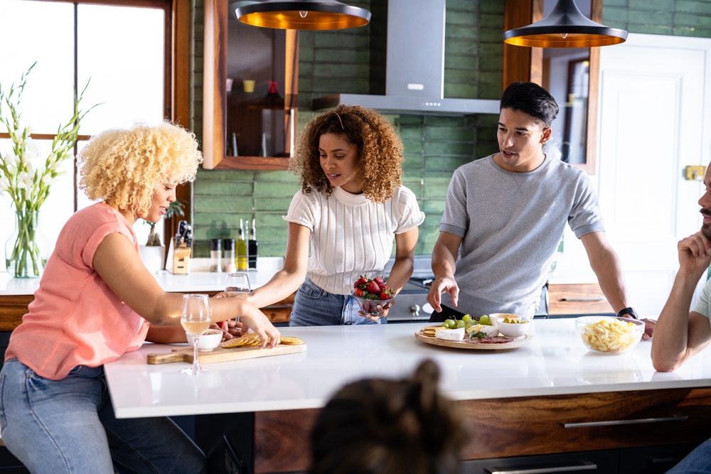 Three people gather around a kitchen island, preparing and sharing snacks and drinks, while chatting. A fourth person, mostly out of frame, sits nearby. The kitchen has green walls and modern decor.