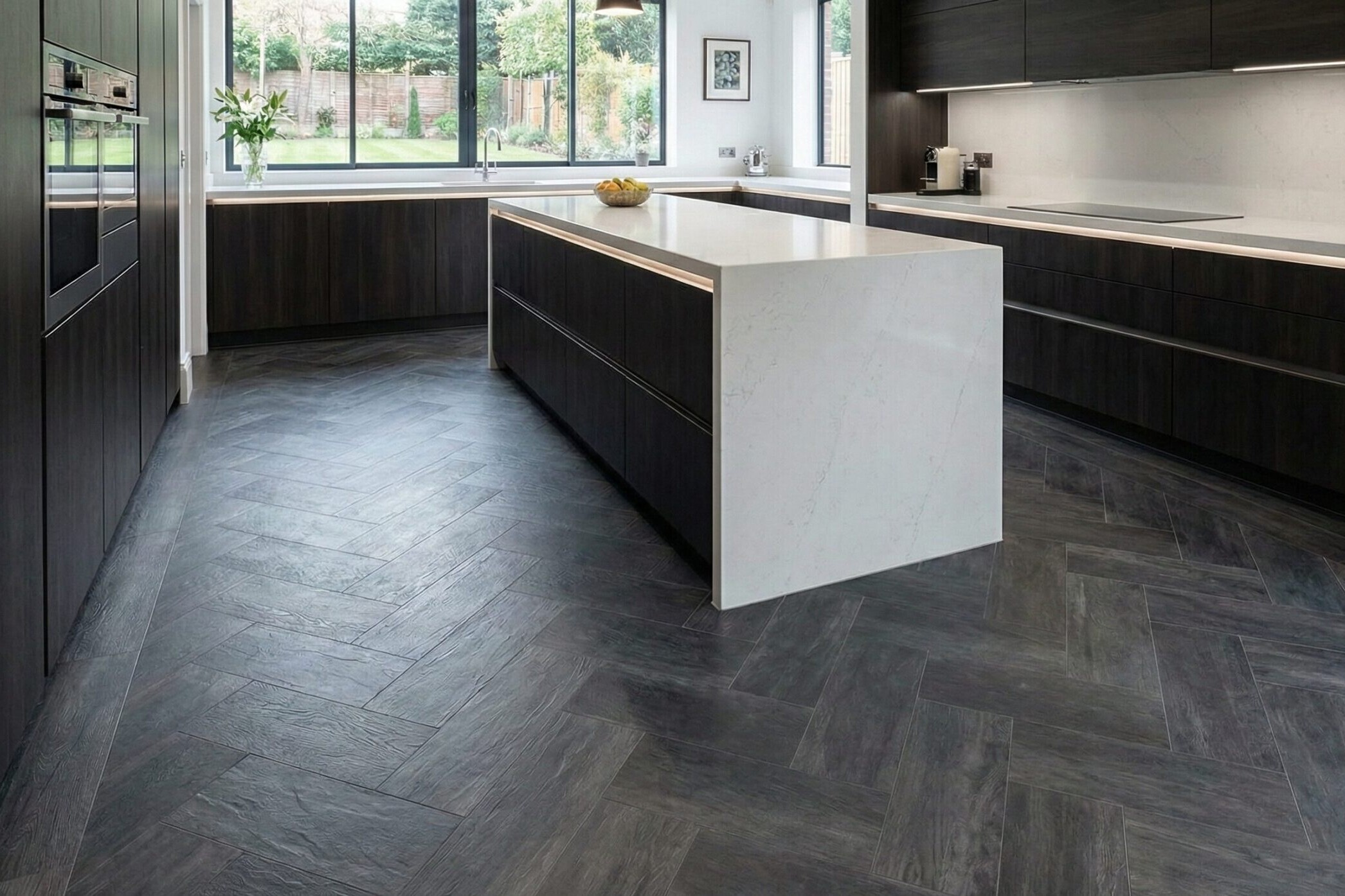 Modern kitchen with dark wood cabinets, a large white marble island, herringbone-patterned dark flooring, built-in appliances, and large windows letting in natural light with a view of the garden.