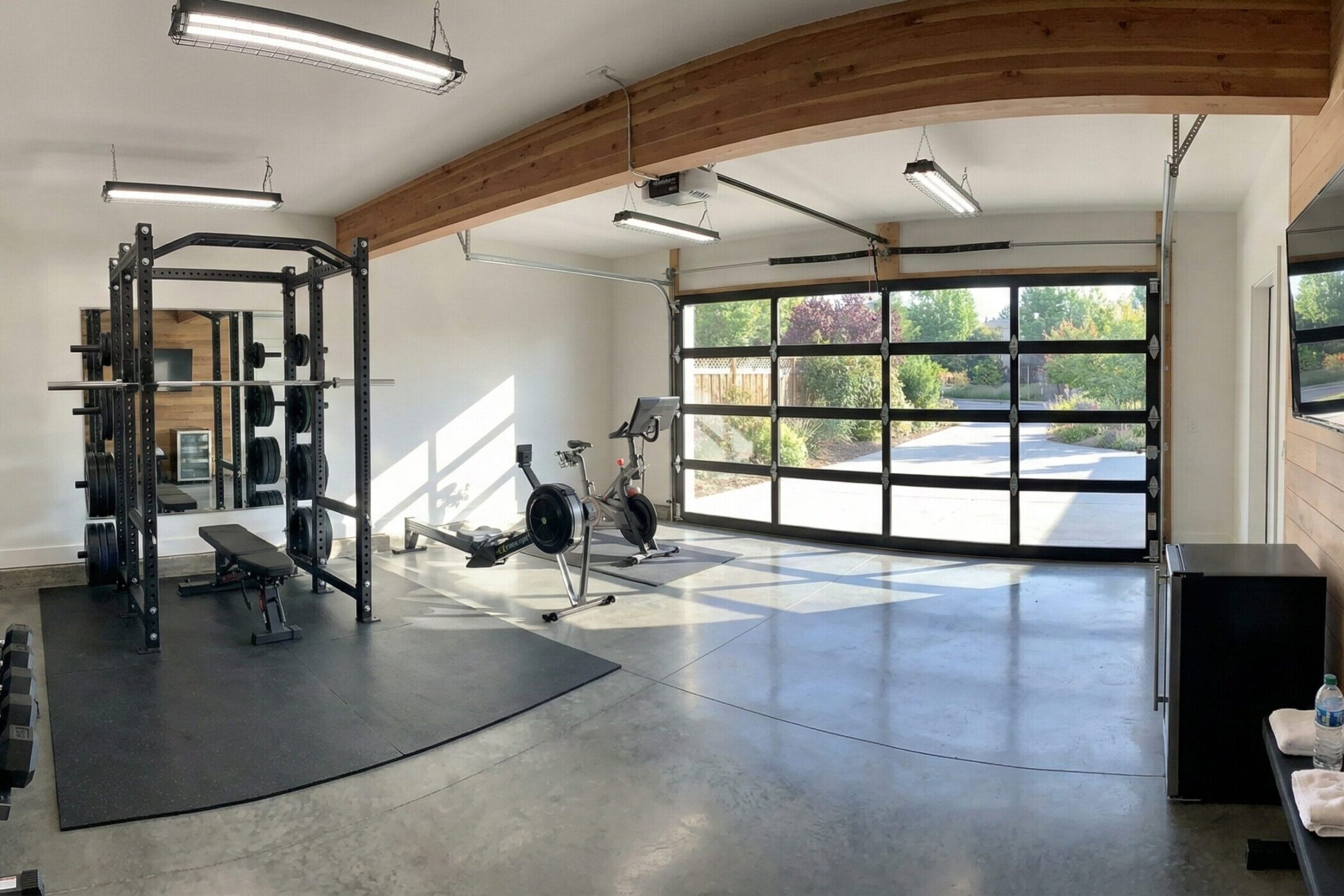 A modern home gym with large windows, dumbbells, a power rack, an exercise bike, rowing machine, wall-mounted TV, and towels, featuring concrete floors and wooden accents with a view of greenery outside.
