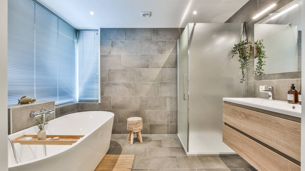 Modern bathroom with a freestanding bathtub, glass-enclosed shower, wood vanity, large gray tiles, window with blinds, hanging plant, and a small wooden stool on a tiled floor.