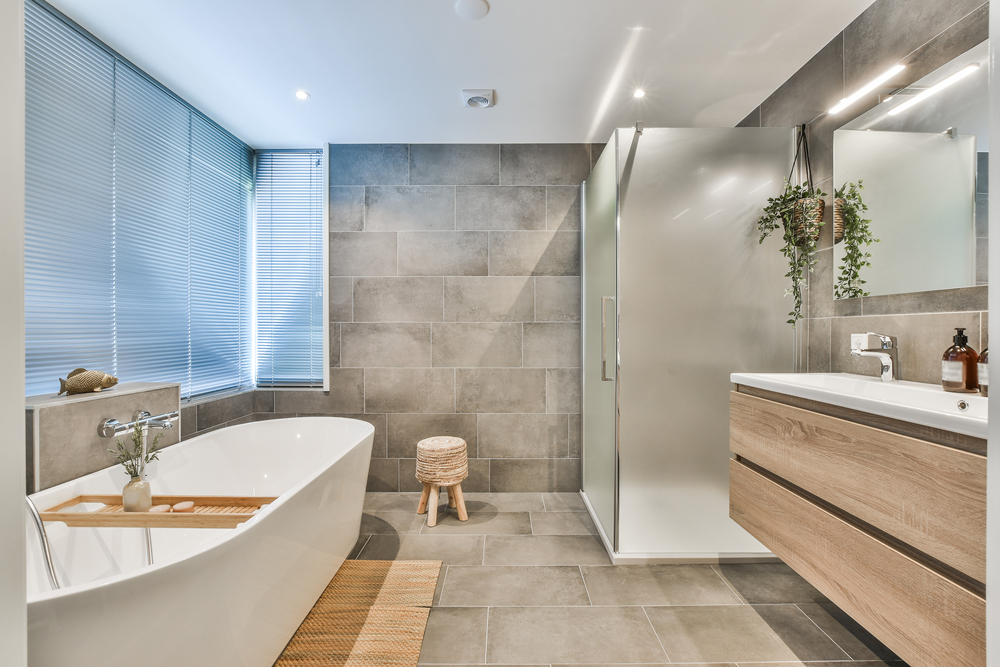 Modern bathroom with a freestanding bathtub, glass-enclosed shower, wood vanity, large gray tiles, window with blinds, hanging plant, and a small wooden stool on a tiled floor.