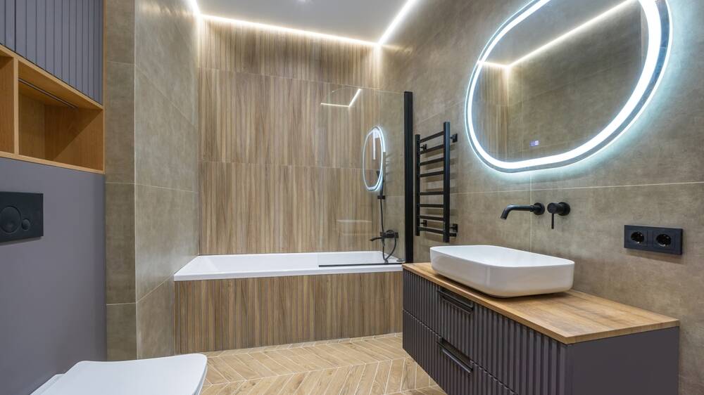 Modern bathroom with wood and beige tiles, a built-in bathtub, a lighted oval mirror, a wall-mounted sink with wooden counter, black fixtures, and a towel rack. The lighting is bright and contemporary.