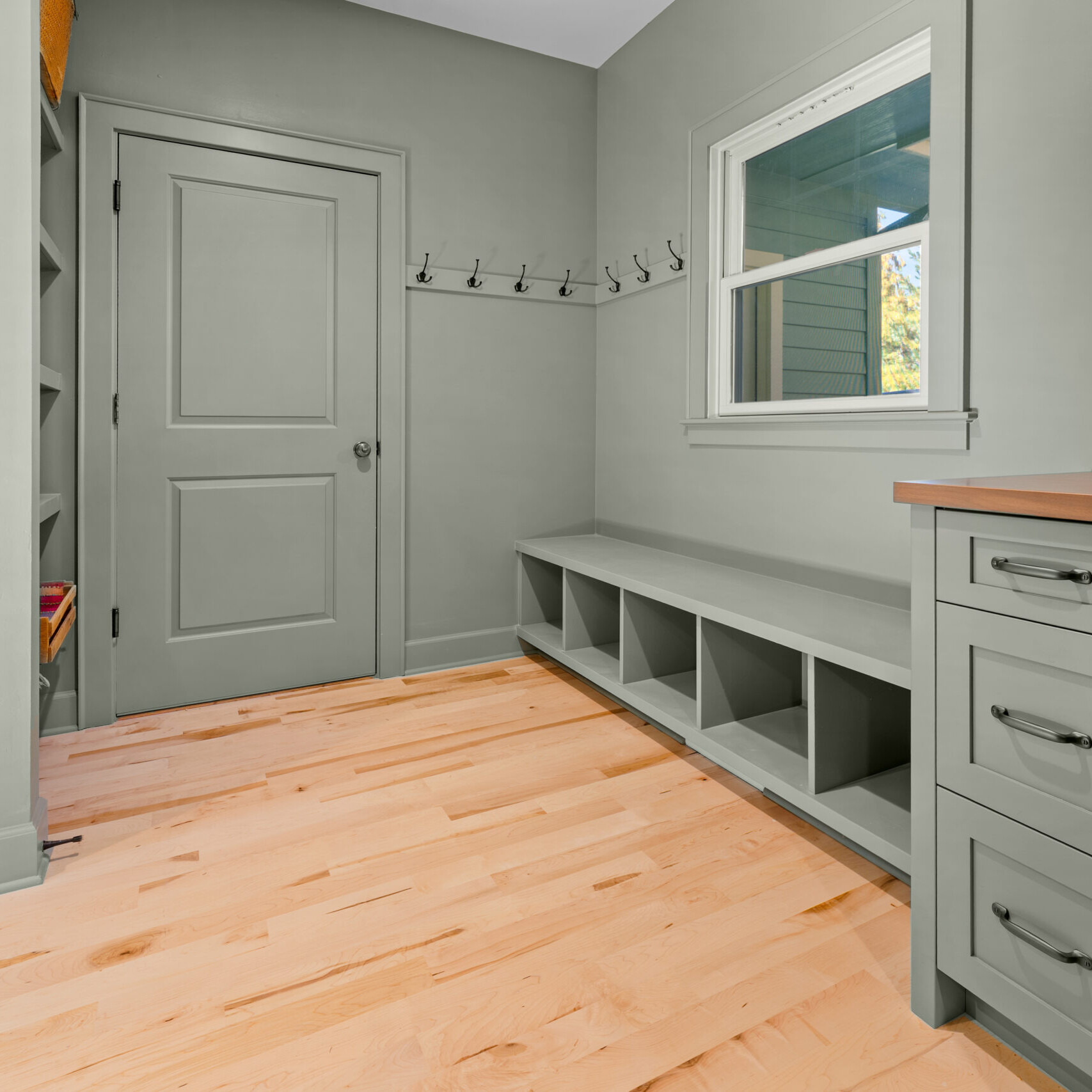 A mudroom with light wood flooring, sage green built-in storage cubbies, cabinets, and hooks on the walls. A window brings in natural light, and a closed door is visible in the background.