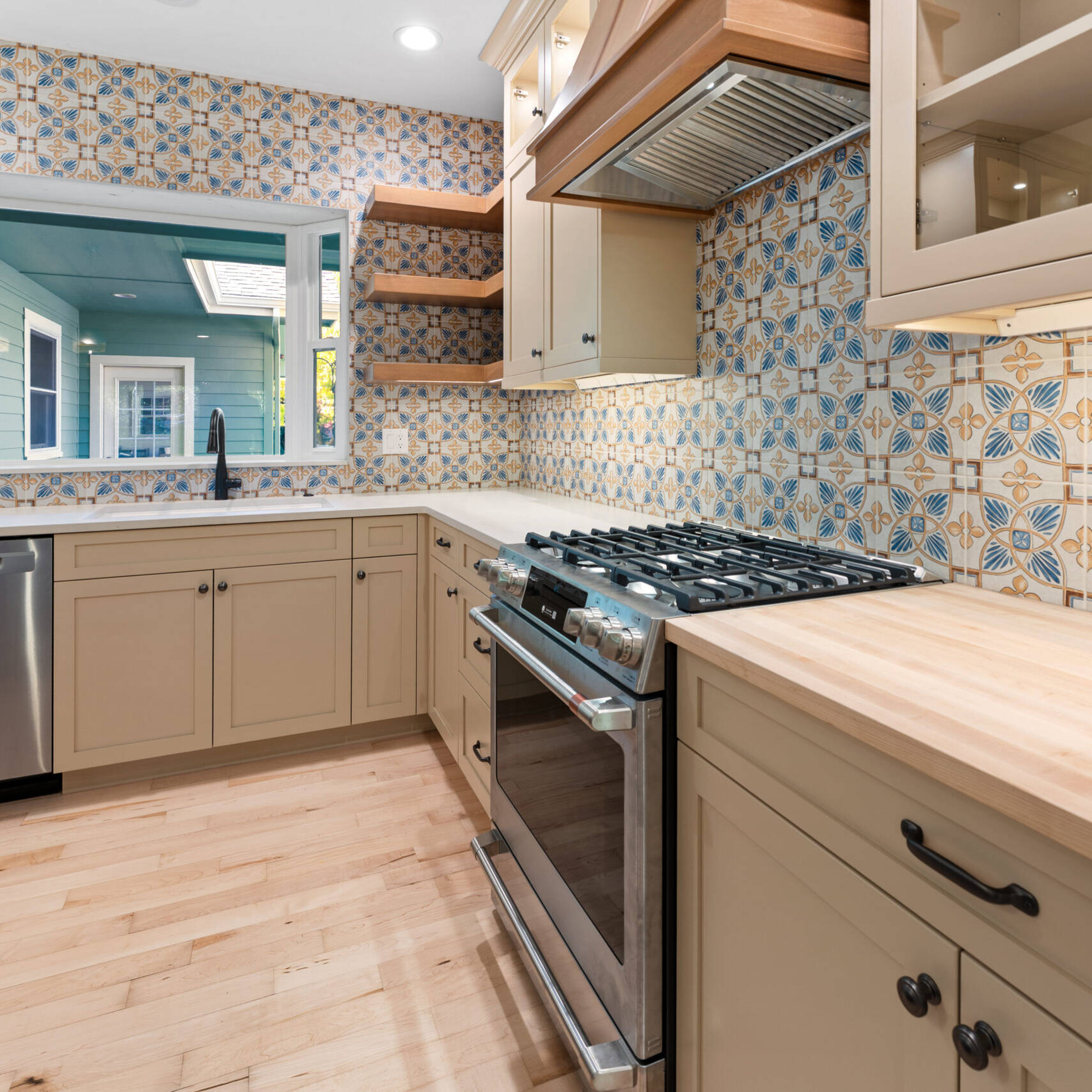 Modern kitchen with patterned blue and beige tile backsplash, light wood cabinets and countertops, a stainless steel stove and dishwasher, open wood shelves, and a large window above the sink.