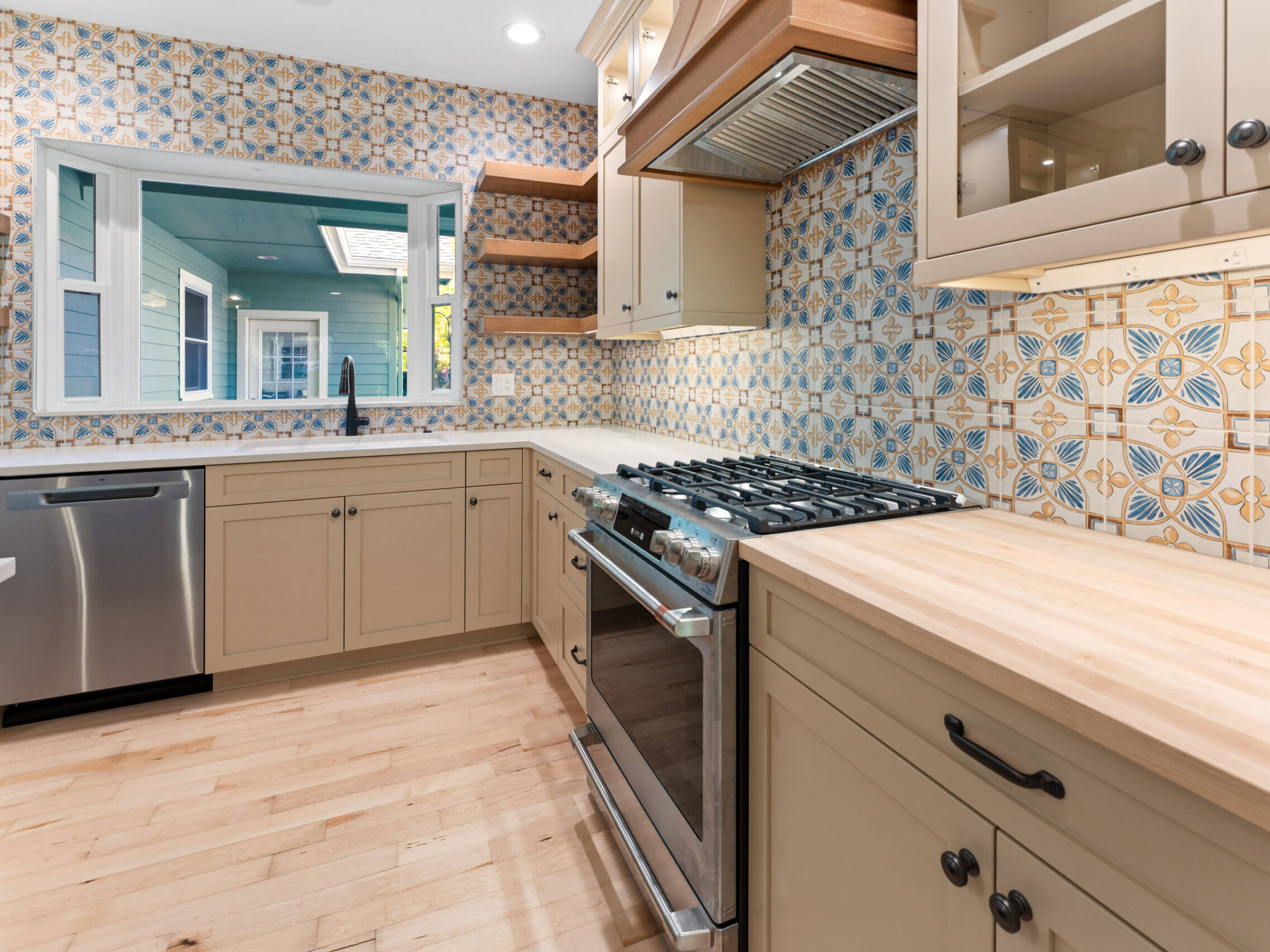 Modern kitchen with patterned blue and beige tile backsplash, light wood cabinets and countertops, a stainless steel stove and dishwasher, open wood shelves, and a large window above the sink.