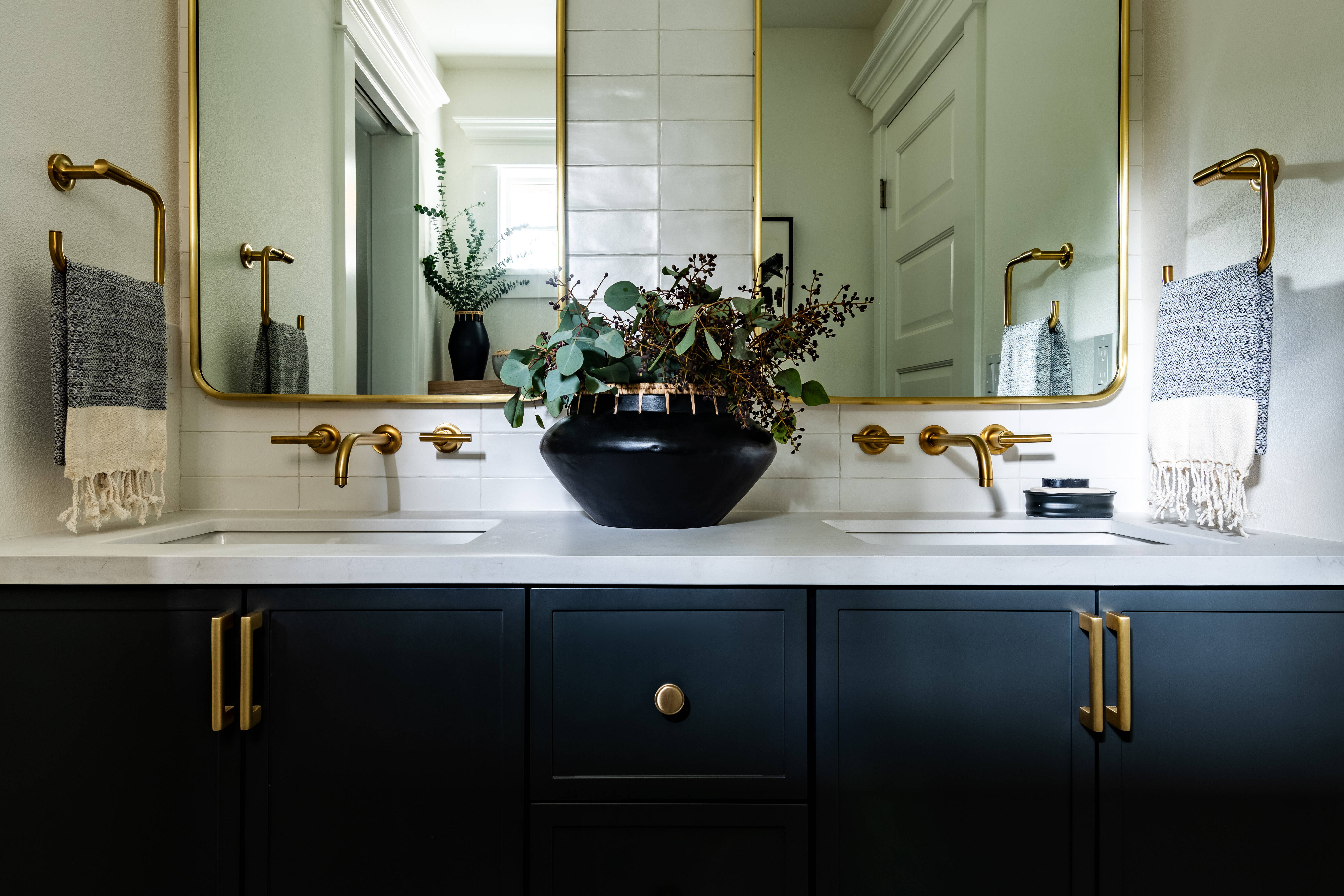 Modern bathroom with double sinks, black cabinets, gold fixtures, two mirrors with gold frames, striped towels, a black vase with greenery, white tile backsplash, and geometric black-and-white floor tiles.