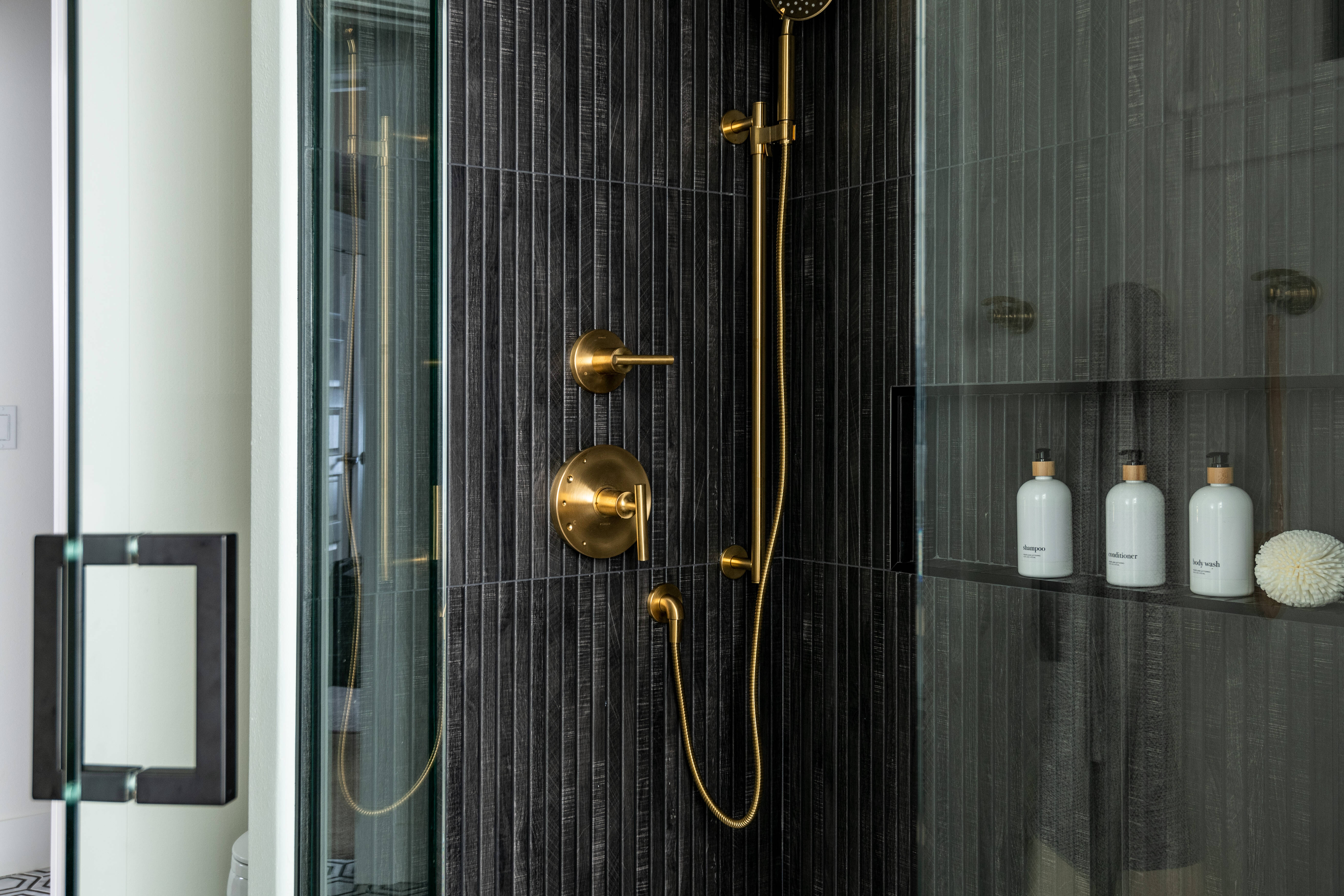 Modern shower with glass doors, black vertical tile walls, gold shower fixtures, a handheld showerhead, and a built-in shelf holding white bottles and a decorative item. The floor features a black and white geometric tile pattern.