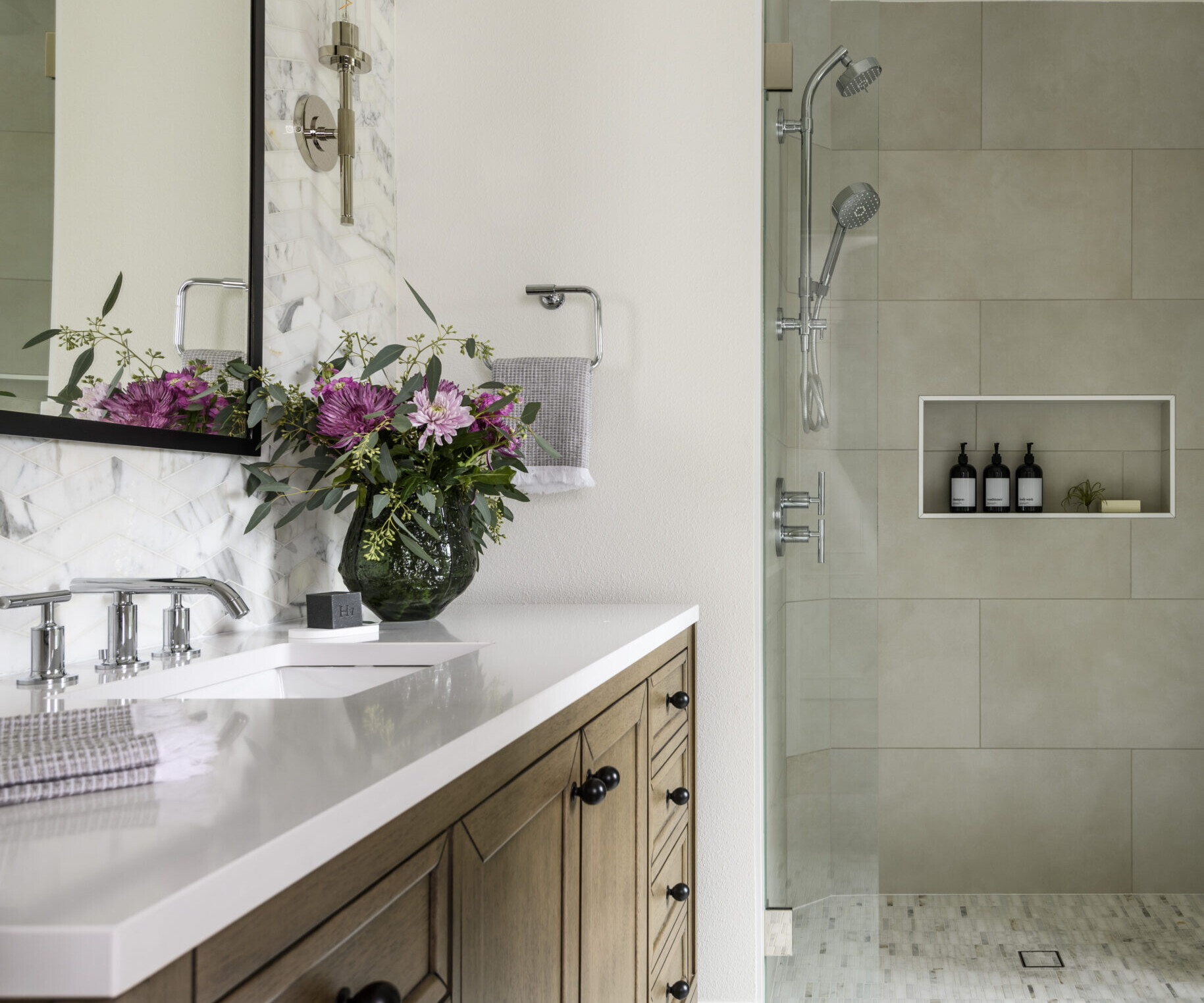 Modern bathroom with a wooden vanity, white countertop, and flowers in a vase. A glass shower with built-in shelves holding toiletries is in the background. The decor features neutral tones and a clean, bright design.