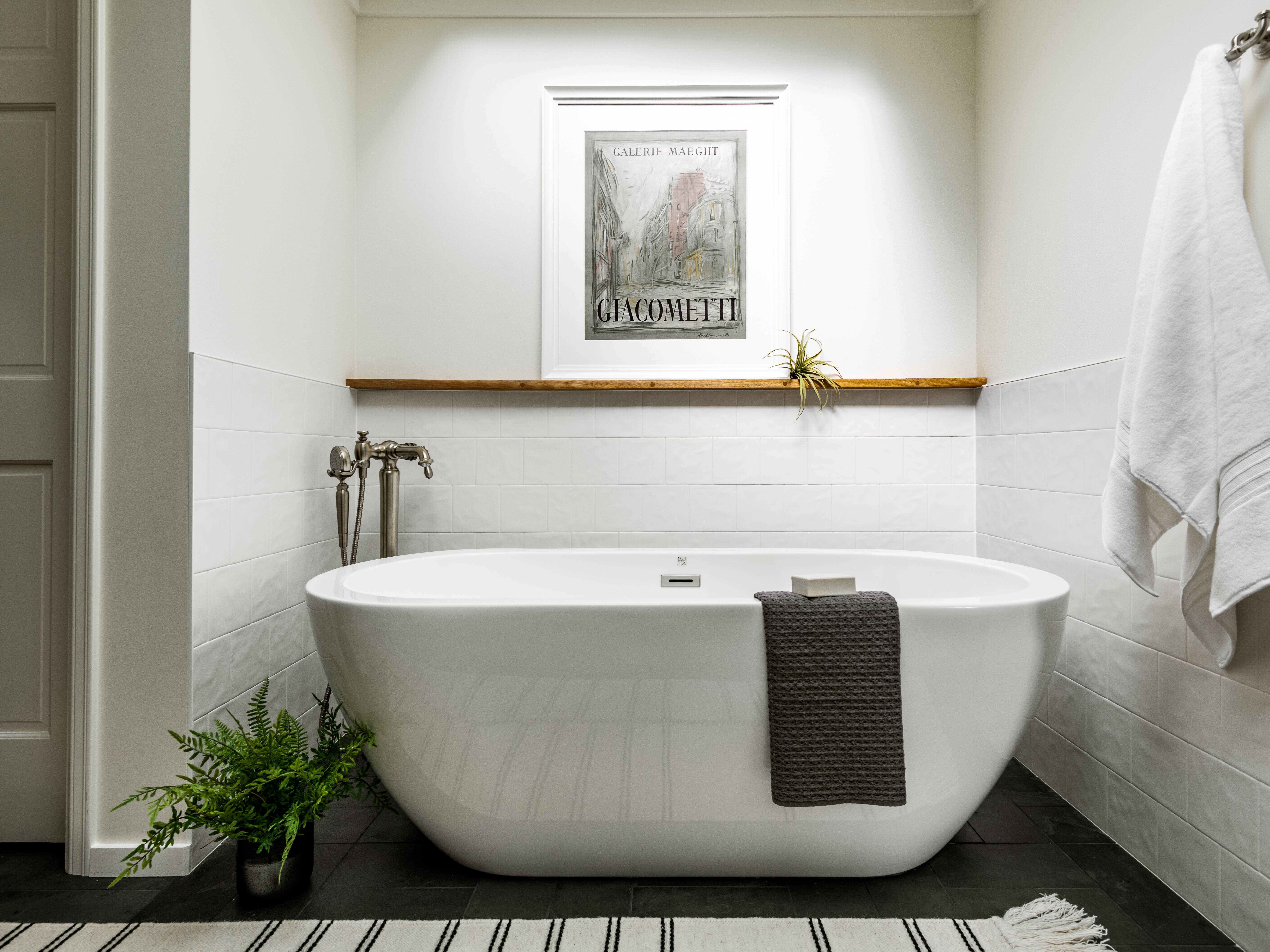 A modern bathroom with a white freestanding tub, a gray towel draped over the side, a fern plant on the floor, a towel hanging on the wall, and a framed artwork above a wooden shelf.