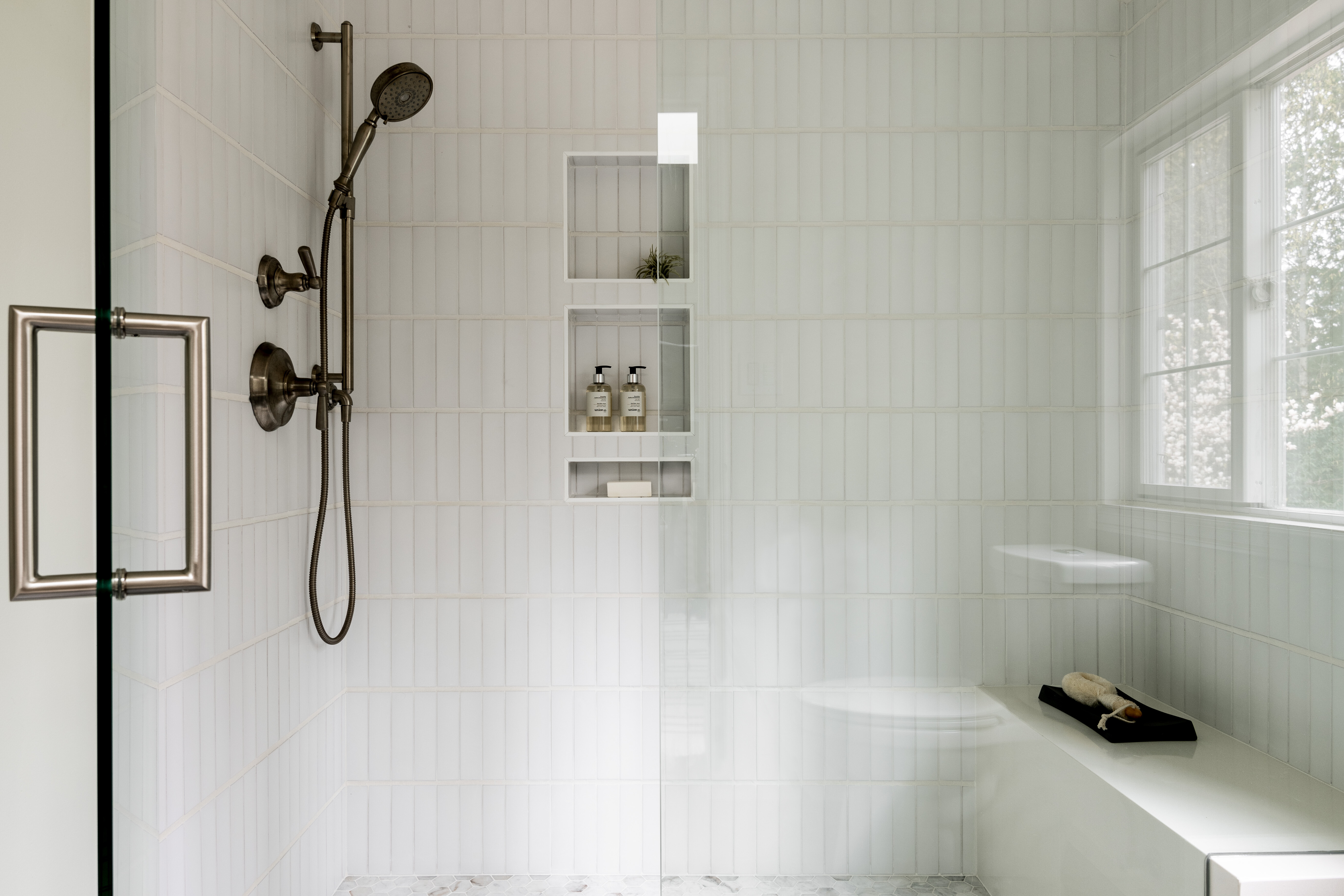Modern shower with glass enclosure, white tile walls, built-in shelving with toiletries, dual shower heads, a handheld shower, and a white bench with a book and towel, set on a patterned shower floor and dark tile outside.