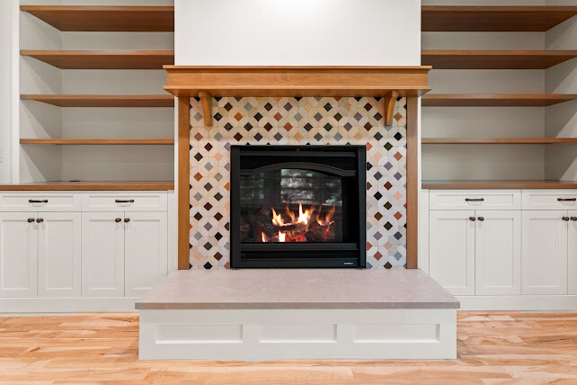 A modern fireplace with a decorative tile surround is centered between built-in white cabinets and wooden shelves, set against a polished wood floor. A fire is burning inside the fireplace.