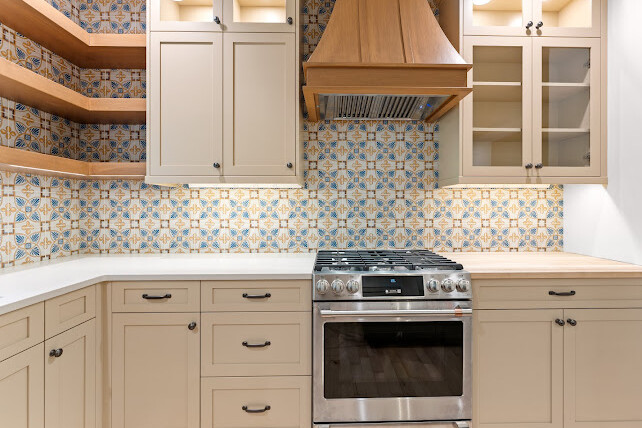Modern kitchen with beige cabinets, a stainless steel gas stove, wooden range hood, open wood shelves, and a colorful patterned tile backsplash. The countertops are white and the floor is light wood.