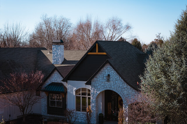 A stone-faced house with dark roof shingles, arched windows, and a chimney, surrounded by bare trees and shrubs, under a clear blue sky.