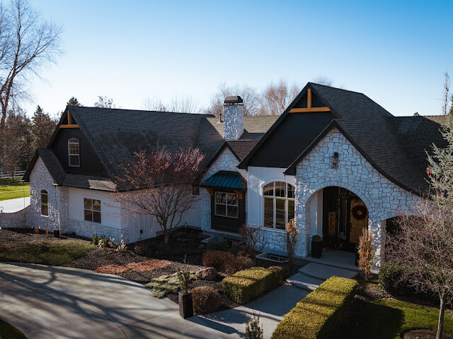 A large white stone house with black roof accents, arched entryway, multiple windows, and a landscaped front yard under a clear blue sky.