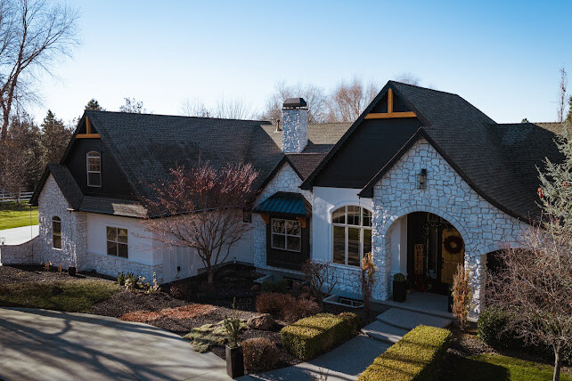 A large white stone house with black roof accents, arched entryway, multiple windows, and a landscaped front yard under a clear blue sky.