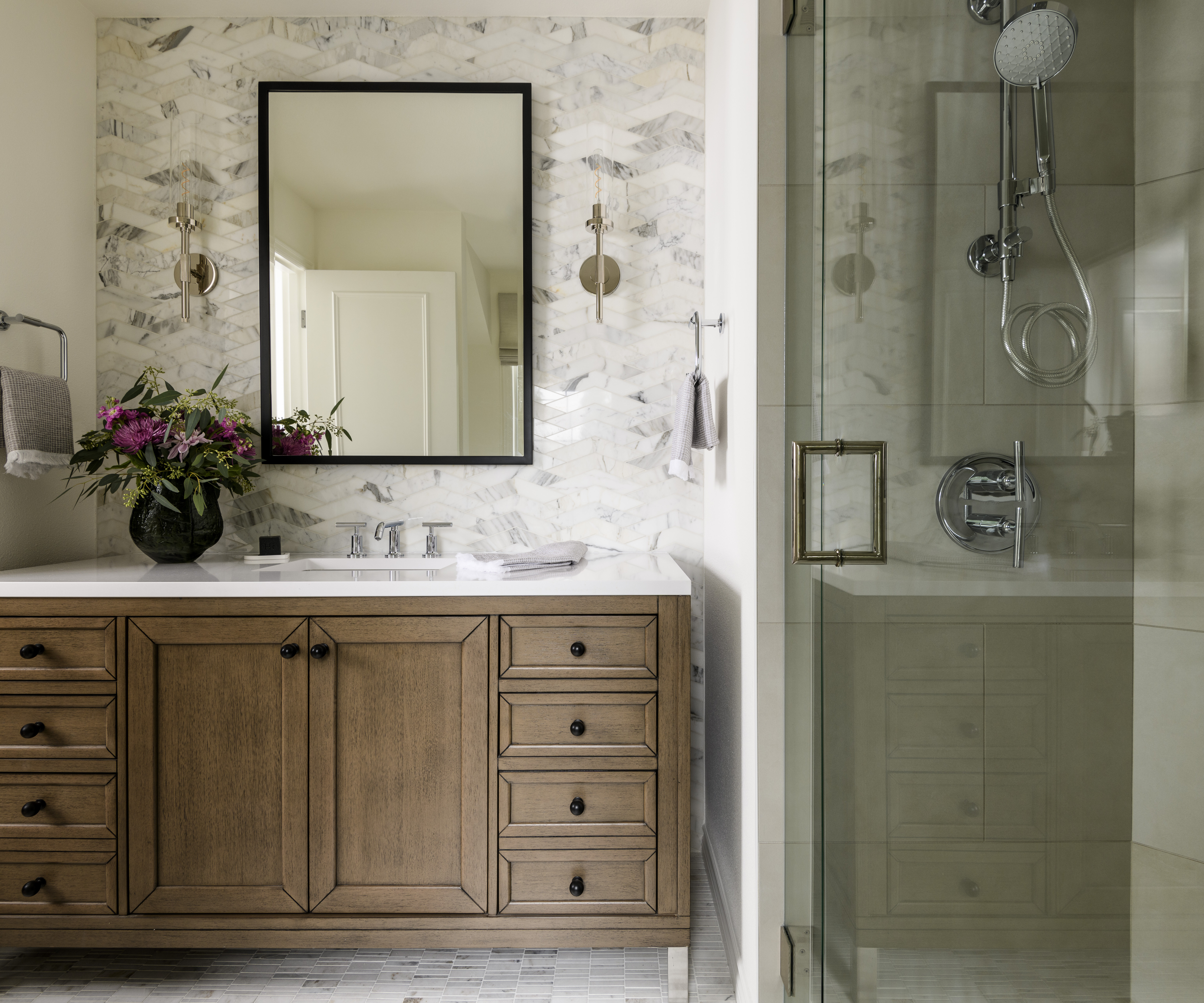 A modern bathroom with a wooden vanity, marble backsplash, rectangular mirror, wall sconces, fresh flowers in a vase, and a glass-enclosed shower featuring chrome fixtures.