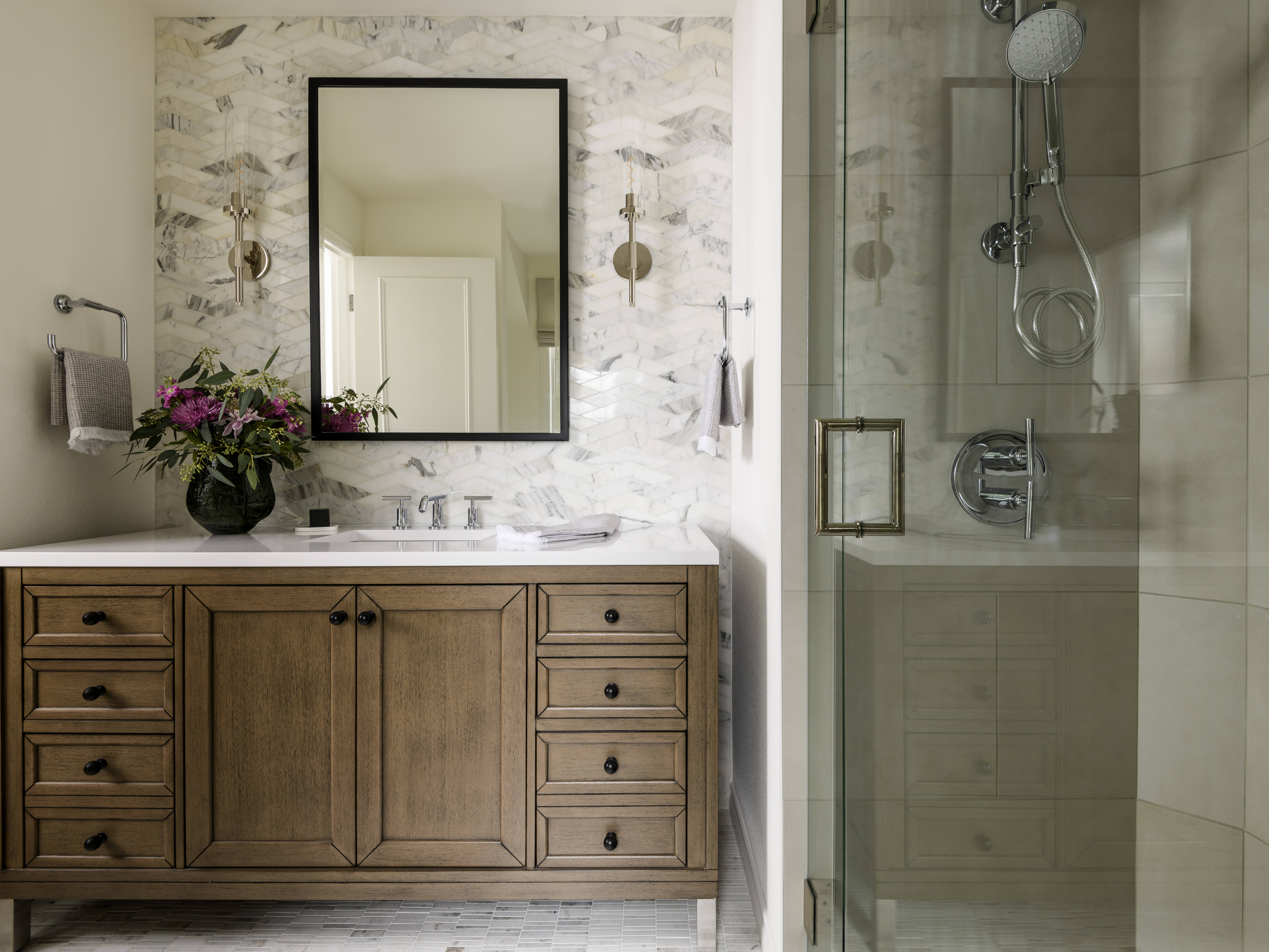 A modern bathroom with a wooden vanity, marble backsplash, rectangular mirror, wall sconces, fresh flowers in a vase, and a glass-enclosed shower featuring chrome fixtures.