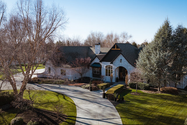 A large house with a dark roof and white exterior stands amid landscaped grounds, featuring a curved driveway, trimmed shrubs, bare trees, and green lawns under a clear blue sky.