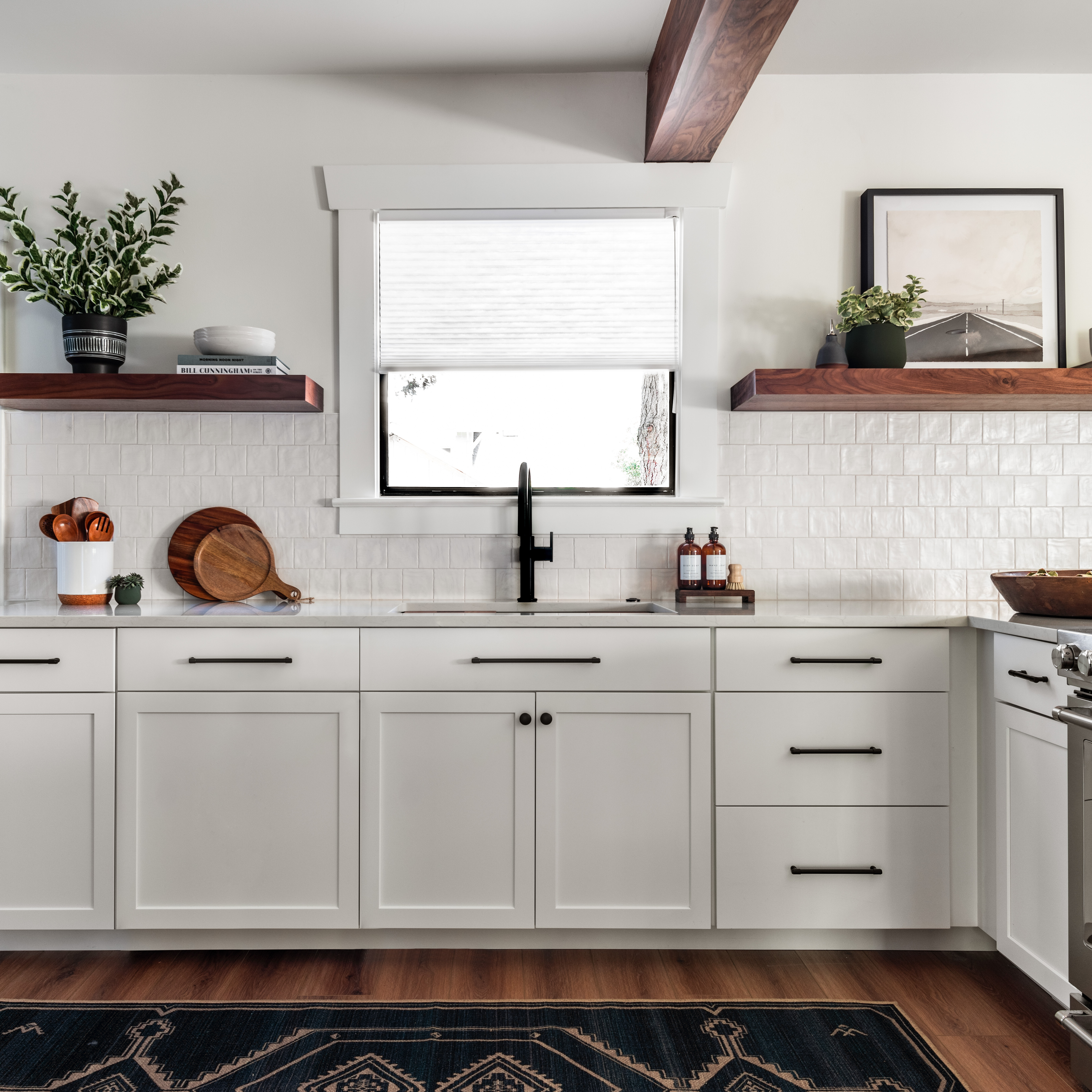 Modern kitchen with white cabinets, black hardware, wood floating shelves, a black faucet, and white tile backsplash. Wooden beams, houseplants, and a pantry with shelves are visible. Natural light fills the space.