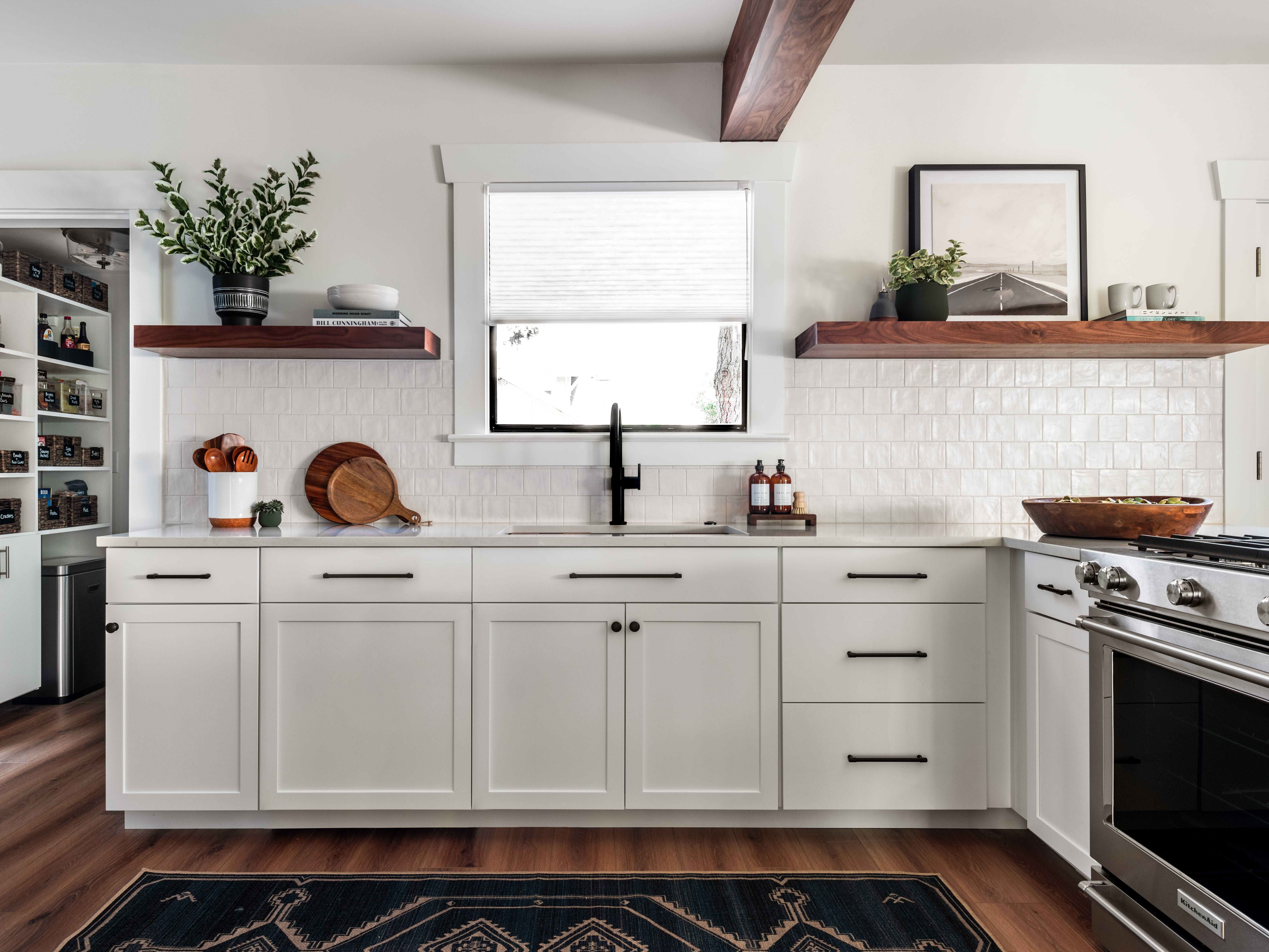 Modern kitchen with white cabinets, black hardware, wood floating shelves, a black faucet, and white tile backsplash. Wooden beams, houseplants, and a pantry with shelves are visible. Natural light fills the space.