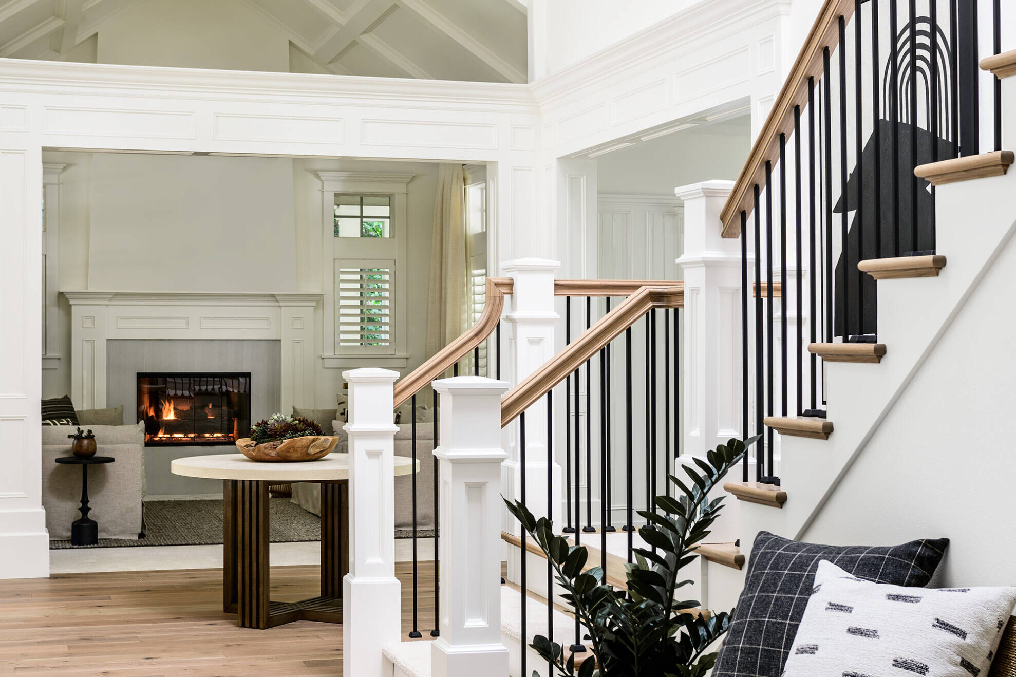A bright, modern entryway with light wood floors, a bench with patterned pillows, a potted plant, black metal stair railings, and a view into a living room with a lit fireplace and neutral décor.