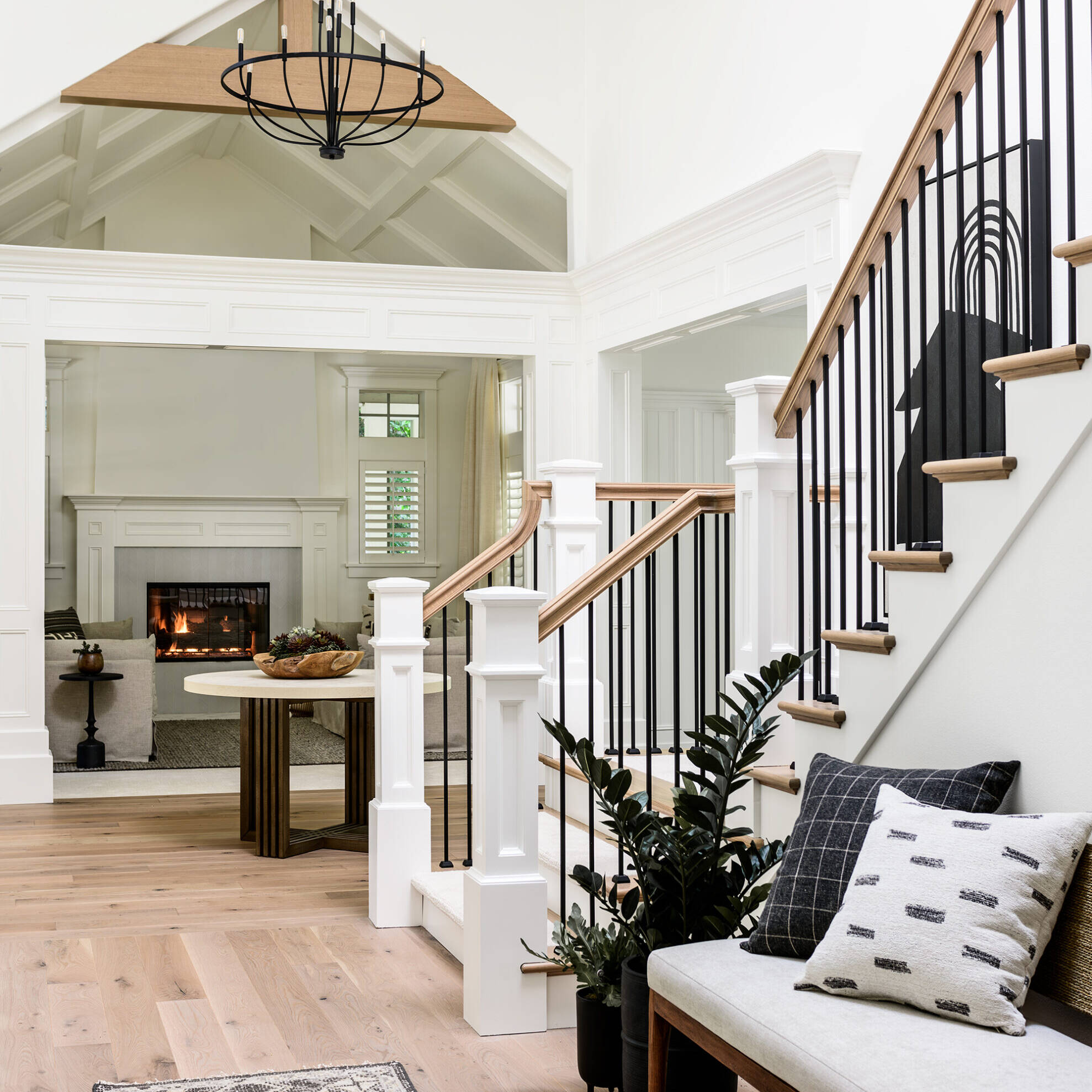 A bright, modern entryway with light wood floors, a bench with patterned pillows, a potted plant, black metal stair railings, and a view into a living room with a lit fireplace and neutral décor.