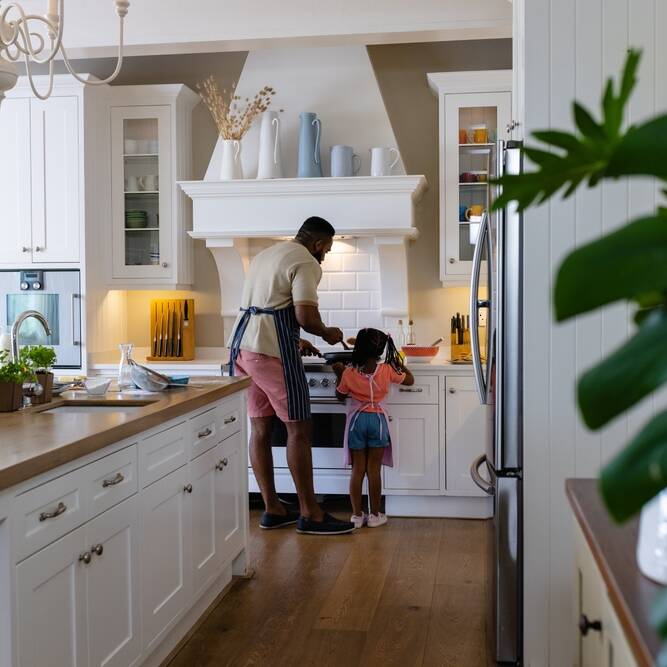 A man and a young girl stand at a stove in a bright, modern kitchen. The man is wearing an apron and cooking, while the girl watches, both seen from behind. The kitchen is tidy with white cabinets and wooden floors.