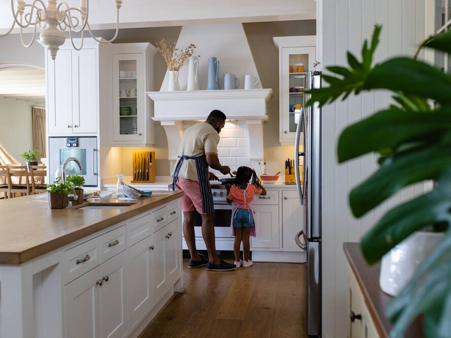 A man and a young girl stand at a stove in a bright, modern kitchen. The man is wearing an apron and cooking, while the girl watches, both seen from behind. The kitchen is tidy with white cabinets and wooden floors.