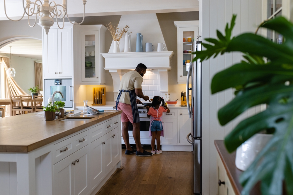 A man and a young girl stand at a stove in a bright, modern kitchen. The man is wearing an apron and cooking, while the girl watches, both seen from behind. The kitchen is tidy with white cabinets and wooden floors.