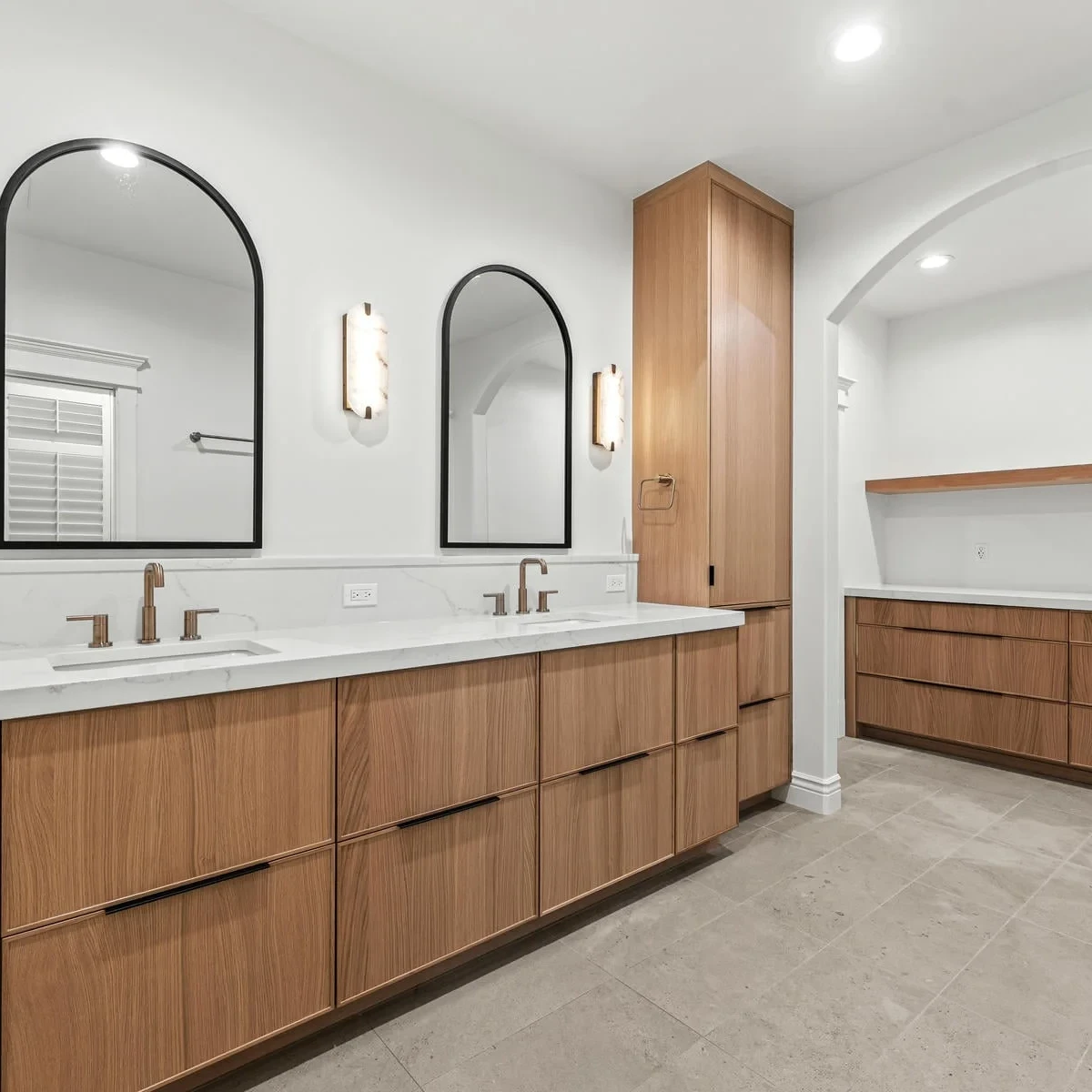 Modern bathroom with double sinks, wood cabinets, arched mirrors, wall sconces, and an additional built-in wood storage area under an arched nook. The design features neutral tones and a clean, minimalist style.