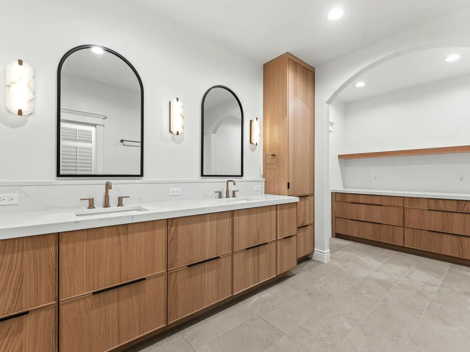 Modern bathroom with double sinks, wood cabinets, arched mirrors, wall sconces, and an additional built-in wood storage area under an arched nook. The design features neutral tones and a clean, minimalist style.
