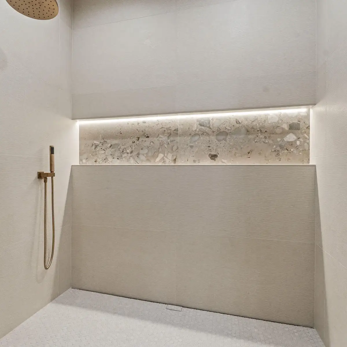 Minimalist walk-in shower with beige tiles, a built-in shelf with stone detail, wall-mounted rainfall showerhead, and a handheld shower. The space is clean and modern, with soft, indirect lighting.