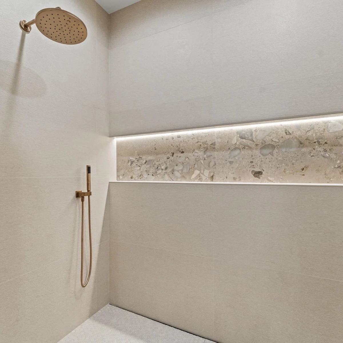 Minimalist walk-in shower with beige tiles, a built-in ledge featuring stone detail, a rainfall showerhead, and a handheld shower, both in a matte gold finish. Recessed lighting highlights the ledge.