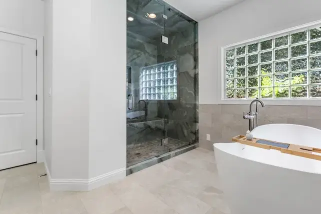 Modern bathroom with a glass-enclosed shower, freestanding white bathtub, large glass block window, and light-colored tile floor and walls. The space is bright and minimalistic.
