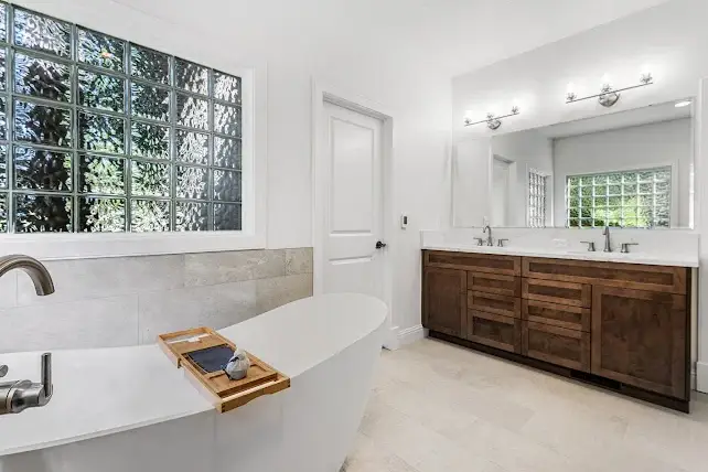 Modern bathroom with a white freestanding tub, a wooden bath tray, a large double-sink vanity with wooden cabinets, glass block window, and a large mirror above the sinks. The space is bright with neutral tones.