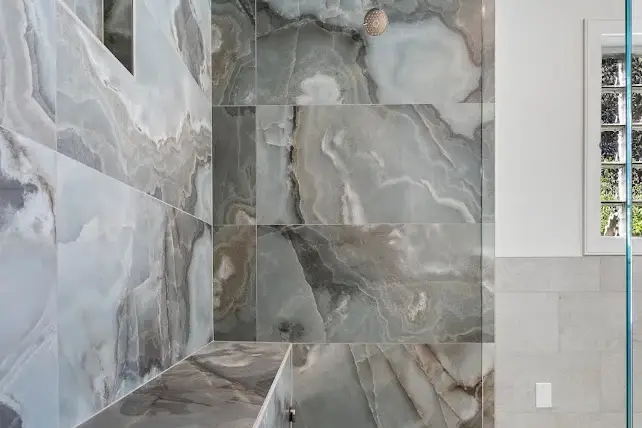 Modern shower with large, glossy gray and white marble-look tiles on the walls and bench, a small dark pebble on the upper wall, and a glass block window on the right side.