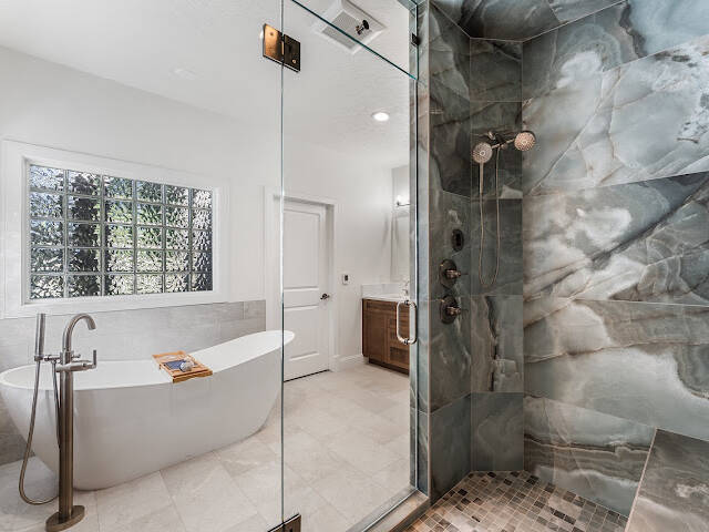 Modern bathroom with a freestanding white tub, a floor-mounted faucet, and a large glass-enclosed shower featuring striking gray and blue stone tiles. Light pours in through a frosted window, brightening the space.