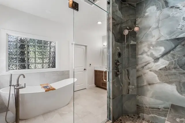 Modern bathroom with a freestanding white bathtub by a large glass block window, and a glass-enclosed shower featuring dark gray and white marble-patterned tiles. Light tile flooring throughout.