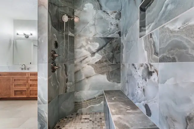 Modern shower with large, gray marble tiles on the walls and bench, dual showerheads, a built-in niche, and a wooden vanity visible in the background.